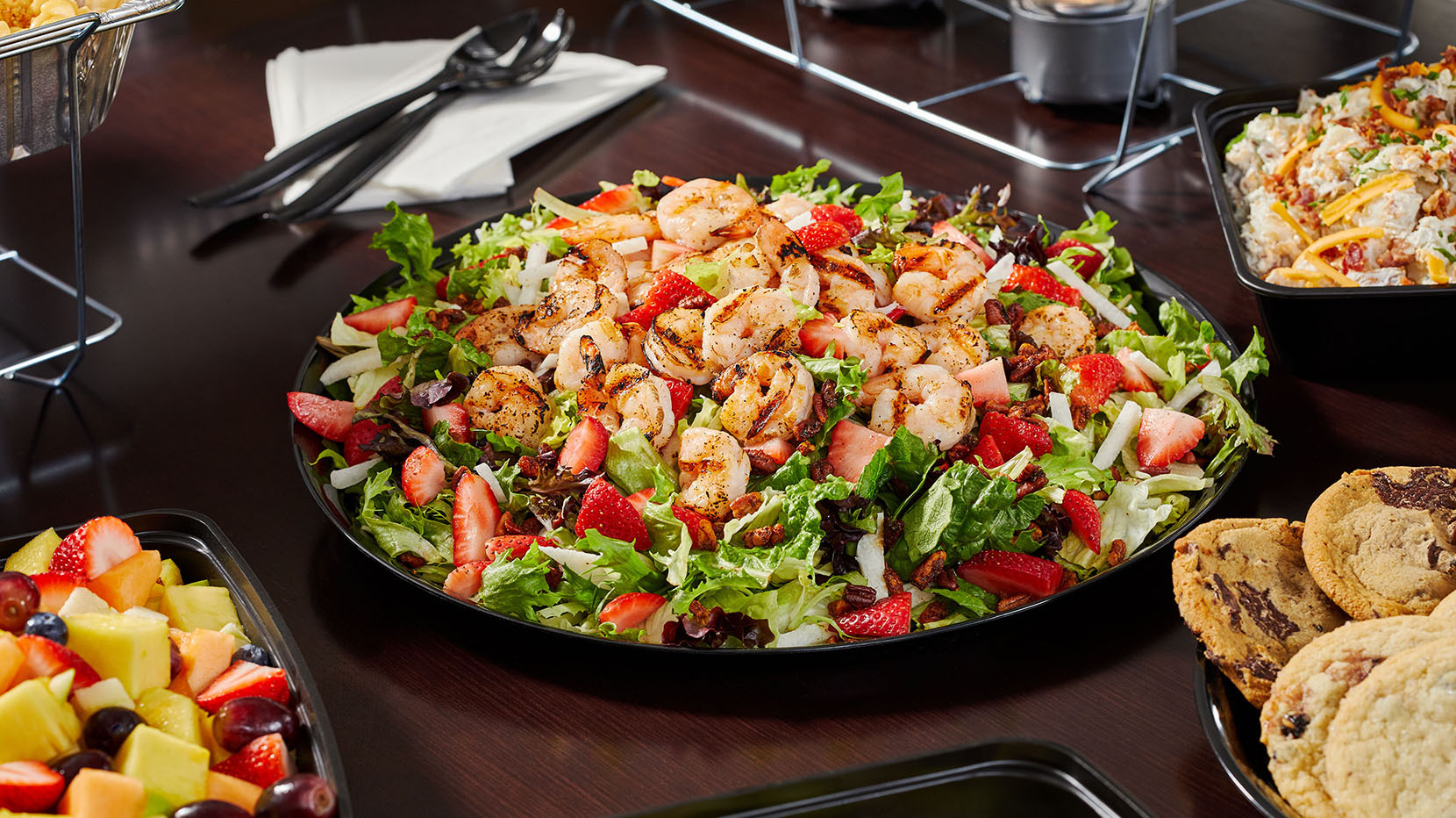 A large share-sized platter of salad surrounded by dishes of fresh fruit, cookies, and more.